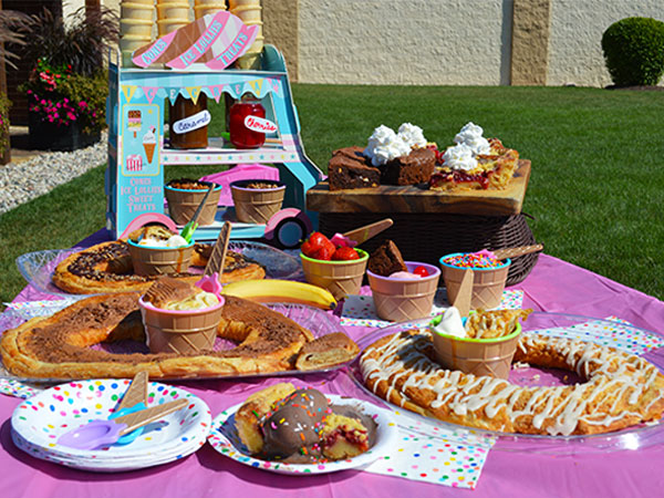 Outdoor picnic spread with three kringles, pie slices, brownies, and ice cream sundaes